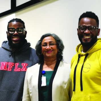 A mature BIPOC female stands flanked by two young black men smiling at the camera.