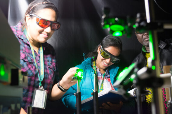 Two women and a man stand with googles infront of a machine with bright green lights