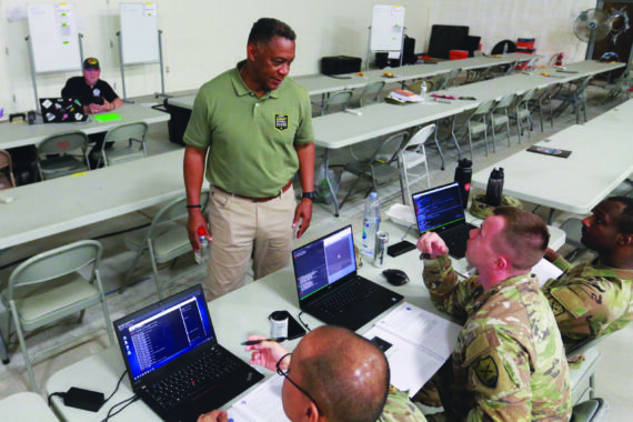 An instructor - a mature black man - stands in front of three military men with computers in a classroom